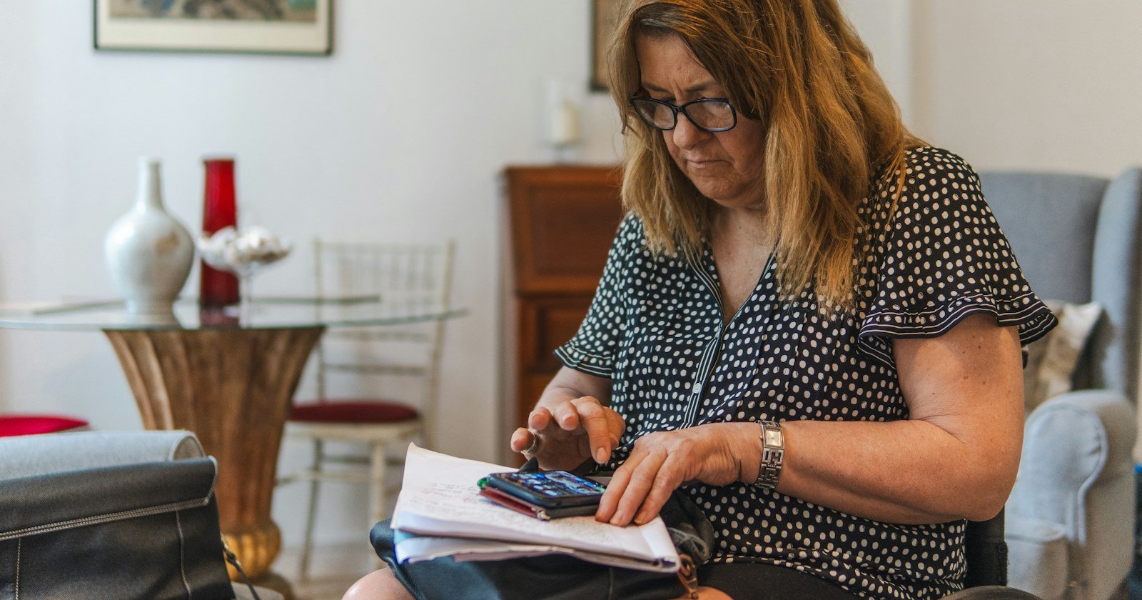 Mulher de óculos conferindo papéis com uma calculadora à mesa de casa